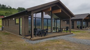 Una cabaña de madera moderna con amplios ventanales, una terraza con asientos exteriores y vistas del paisaje circundante.