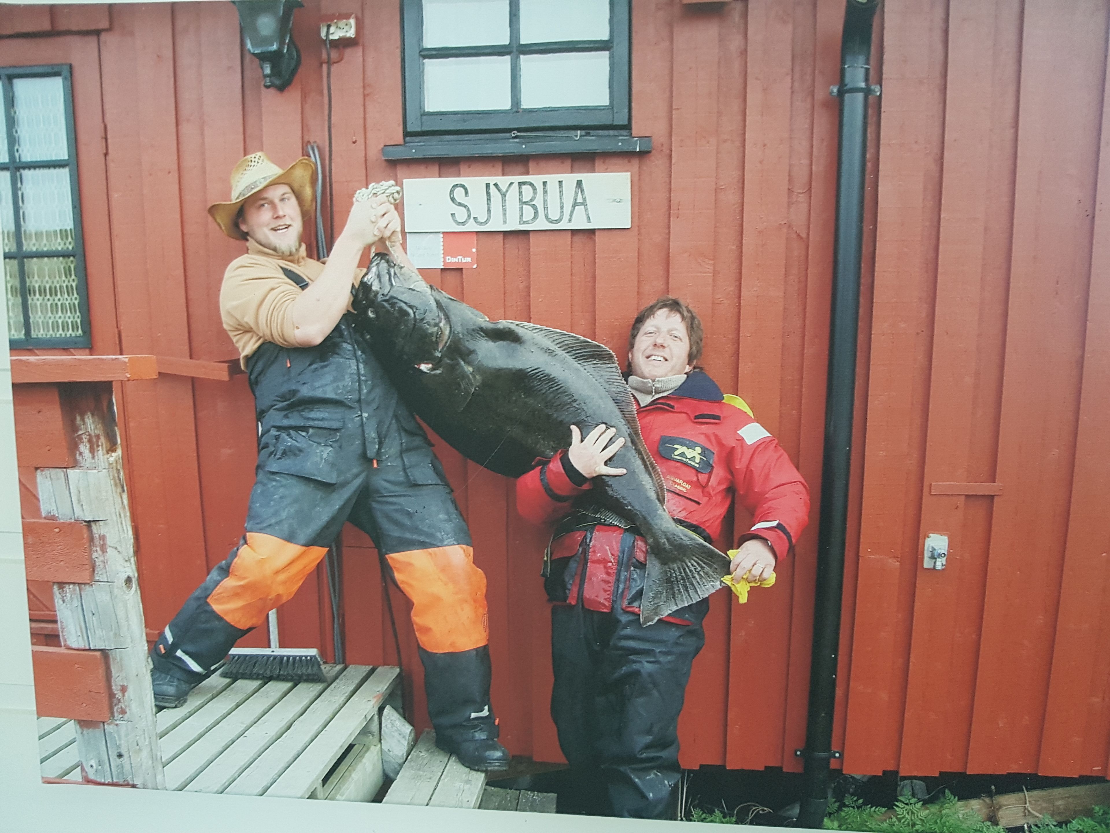 Two people holding a large fish in front of a red building with a sign that reads "SJYBUA".