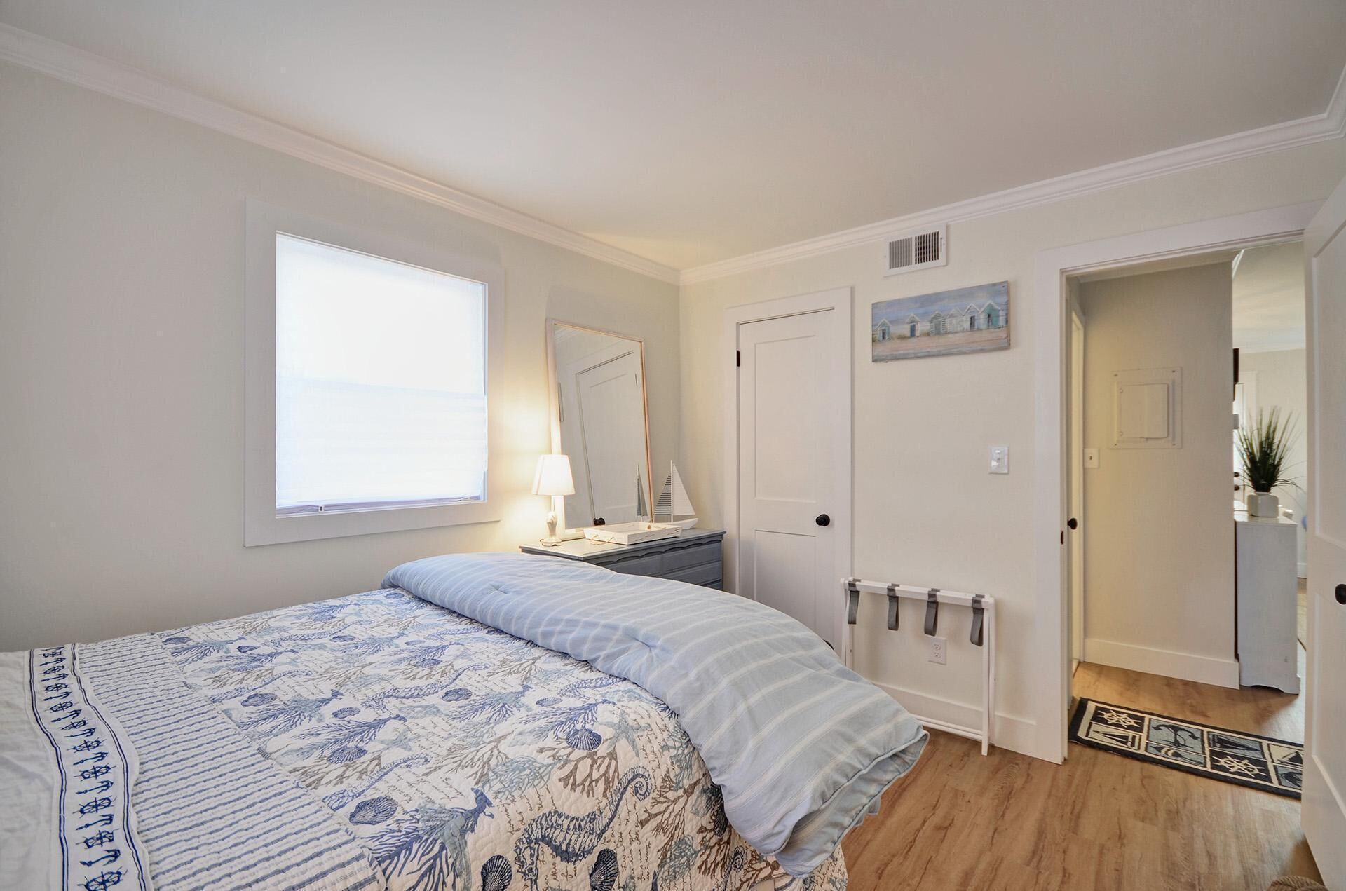 A bedroom with a bed, a nightstand, a lamp, a dresser, a door, and a small painting on the wall.