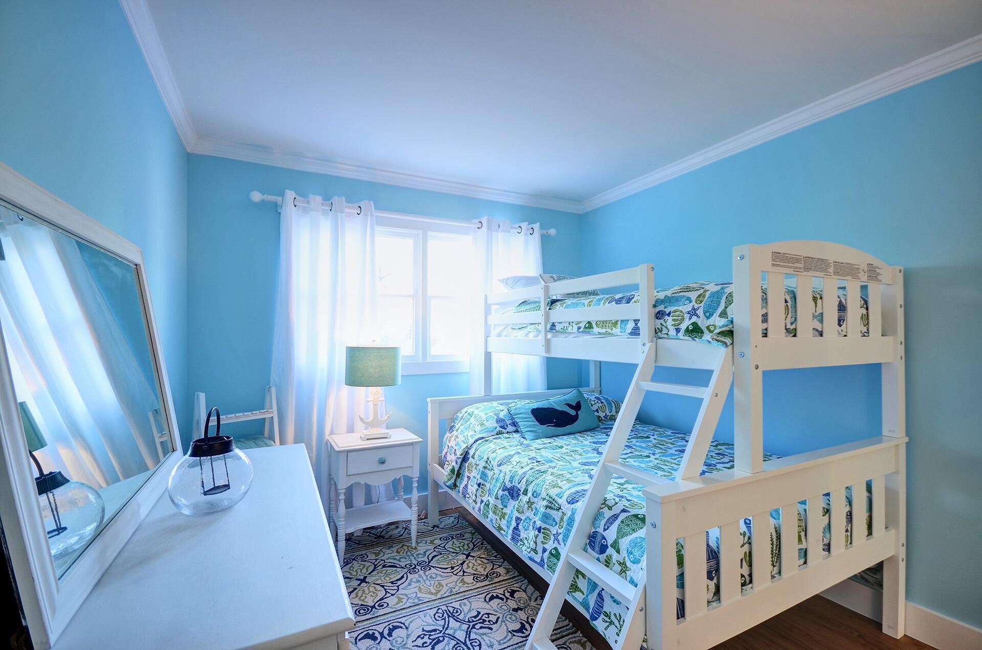 A child's bedroom with a bunk bed, a bedside table, a lamp, and a window with curtains.