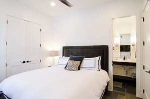 Un dormitorio con una cama grande, cabecera oscura, dos lámparas de noche, una puerta blanca y un baño con espejo y lavamanos.