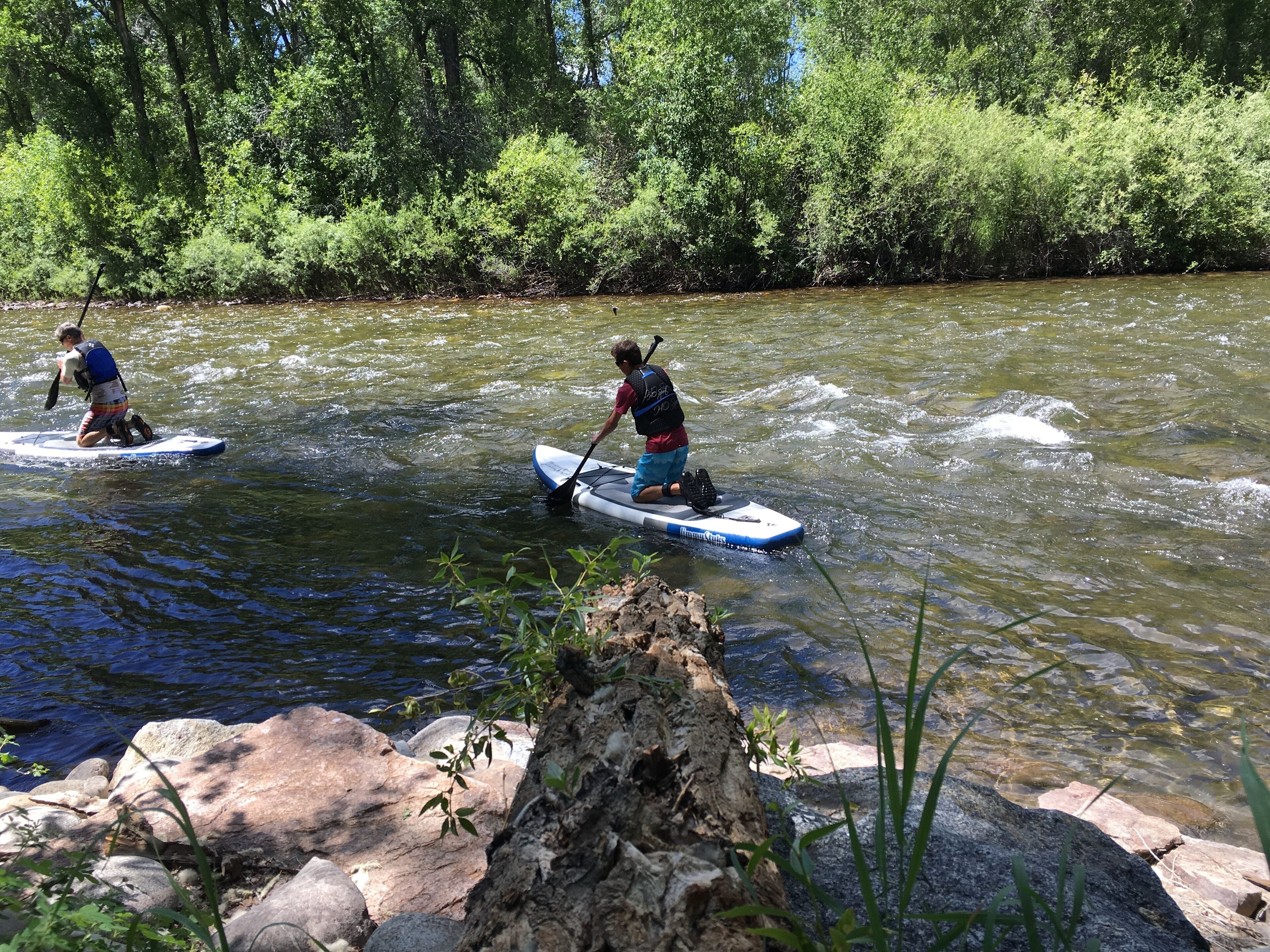 Zwei Personen stehen auf Paddleboards auf einem Fluss, umgeben von Felsen und Grünpflanzen.