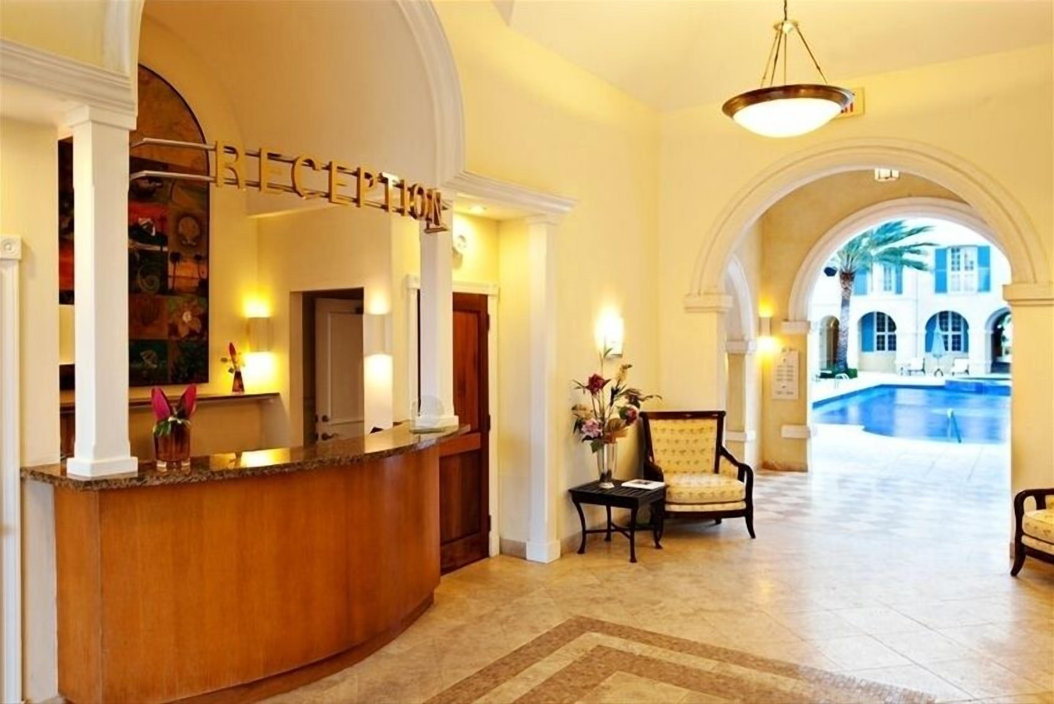Un hall d’hôtel comprenant un comptoir d’accueil, un espace détente avec des fauteuils et une vue sur une piscine, visible à travers une arche.