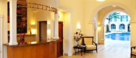 Un hall d’hôtel comprenant un comptoir d’accueil, un espace détente avec des fauteuils et une vue sur une piscine, visible à travers une arche.