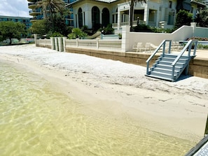 A beachfront hotel with clear shallow waters, a sandy beach, and a staircase leading to a balcony.