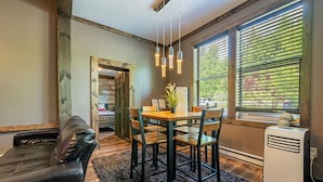 A cozy room with a wooden dining table, four chairs, a black leather sofa, and a large window with blinds.