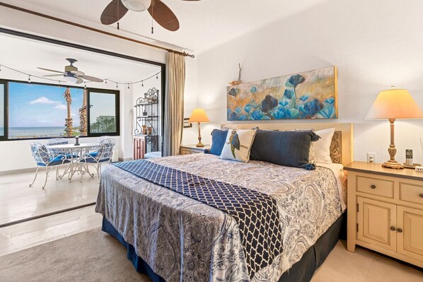 A bedroom with a large bed, a desk, and a chair, a ceiling fan, and a view of the ocean.