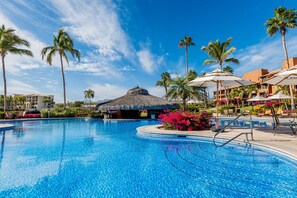 A clear blue pool with lounge chairs and umbrellas, surrounded by palm trees and a thatched-roof structure.