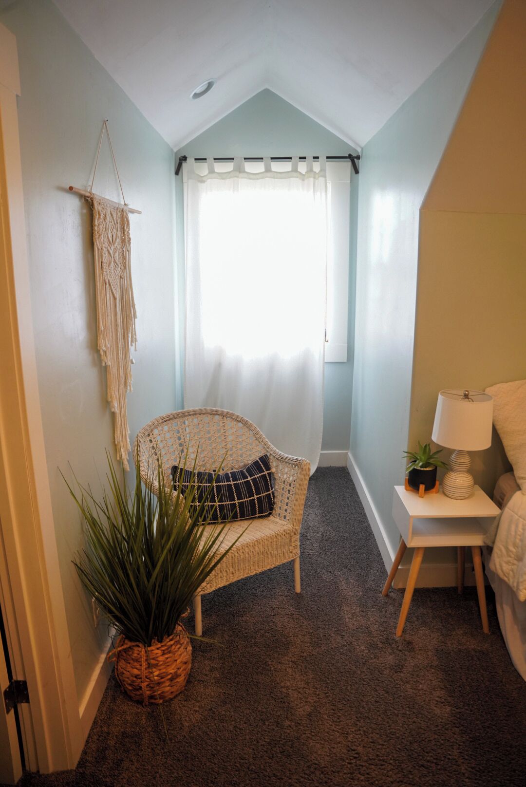 A cozy room with a wicker chair, a small table with a lamp, a potted plant, and a window with sheer curtains.