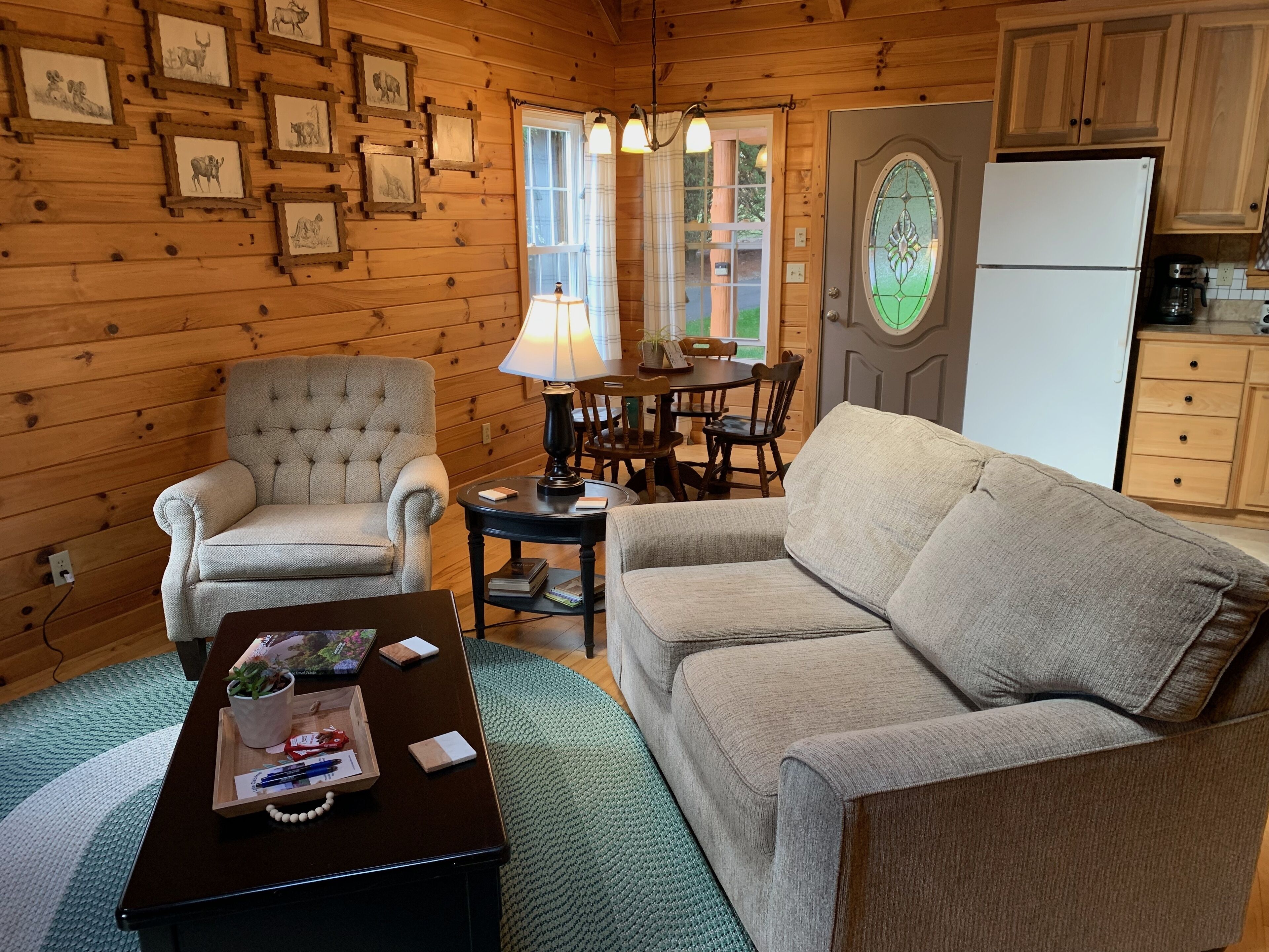 A cozy cabin interior with a wooden wall, a dining table with chairs, a sofa, a coffee table, and a refrigerator.