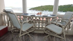 Un balcone con un tavolo apparecchiato per due, con vista su un tratto d'acqua, imbarcazioni e una collina verde sullo sfondo.