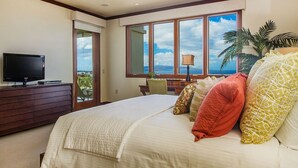 Quarto com cama grande, televisão em cômoda, escrivaninha com abajur e vista para o mar por grandes janelas.
