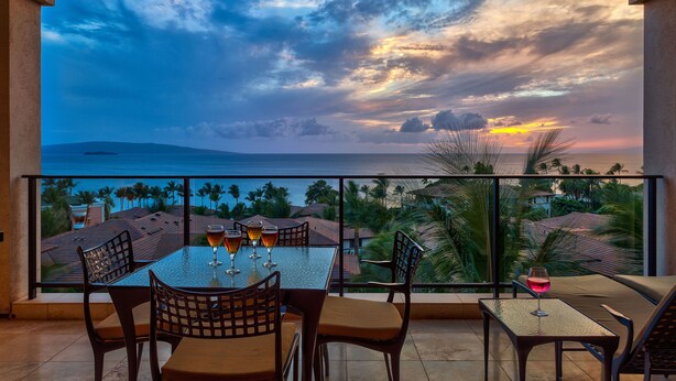 Uma varanda com mesa e cadeiras, com vista para o pôr do sol, palmeiras e uma ilha ao longe.