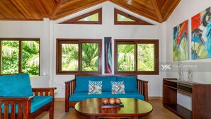 A room with a wooden ceiling, a sofa with teal cushions, a coffee table, and large windows with a tropical view.