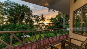 A balcony with a view of a sunset, surrounded by lush greenery and palm trees.