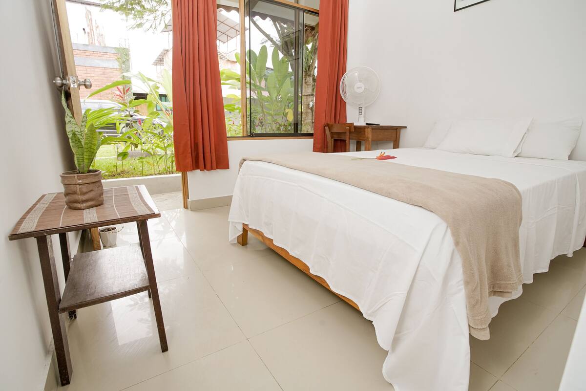 Une chambre à coucher avec un lit, une table de chevet, un ventilateur et une fenêtre avec des rideaux.