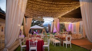 Un salón de bodas al aire libre con mesas y sillas blancas, cortinas drapeadas e iluminación con luces colgantes.