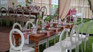 Un área de comedor al aire libre con mesas de madera y sillas blancas, decorada con manteles rosa y cristalería.