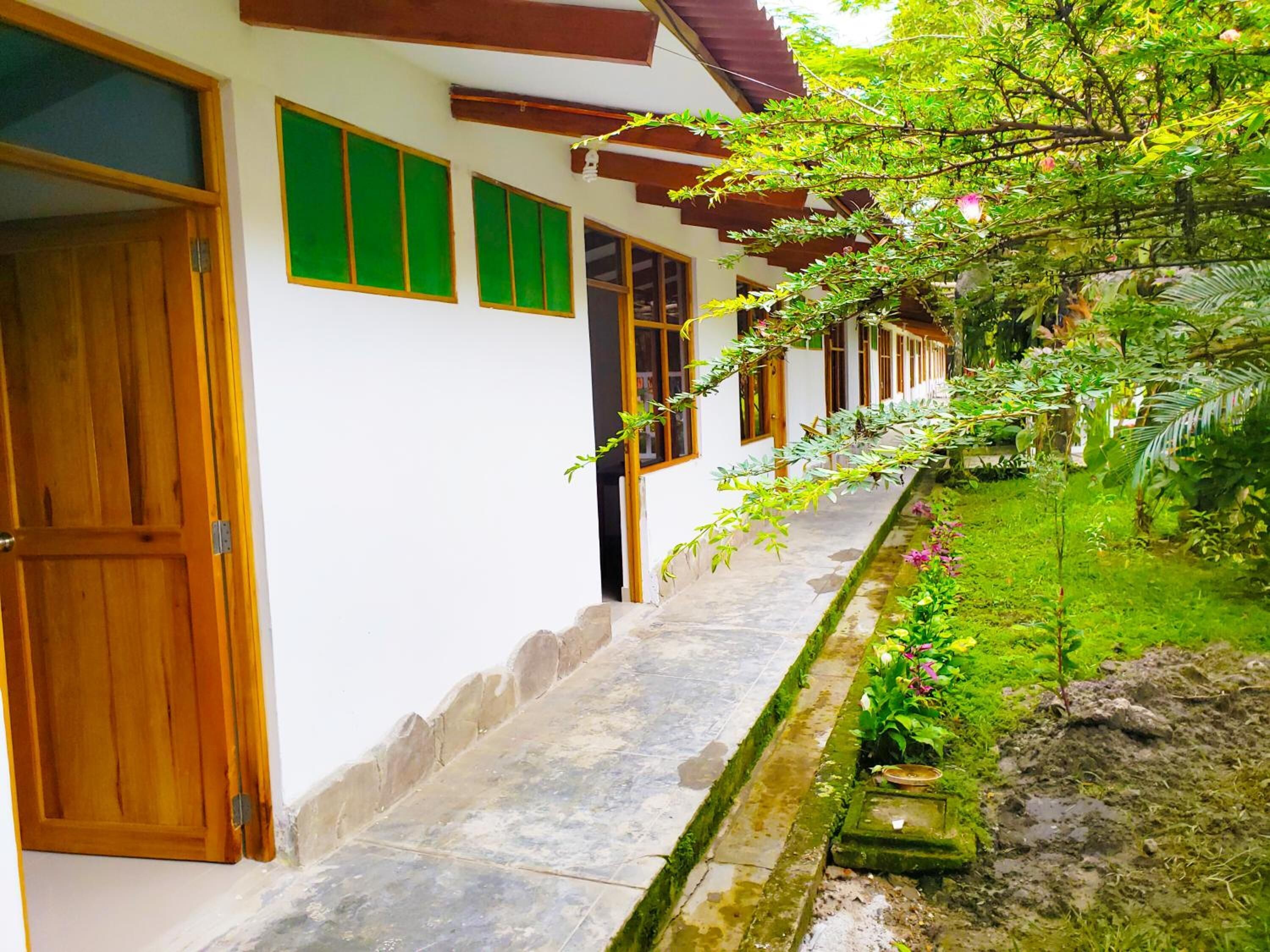 A building with a long, narrow corridor with wooden doors and windows, surrounded by lush greenery and a small garden area.