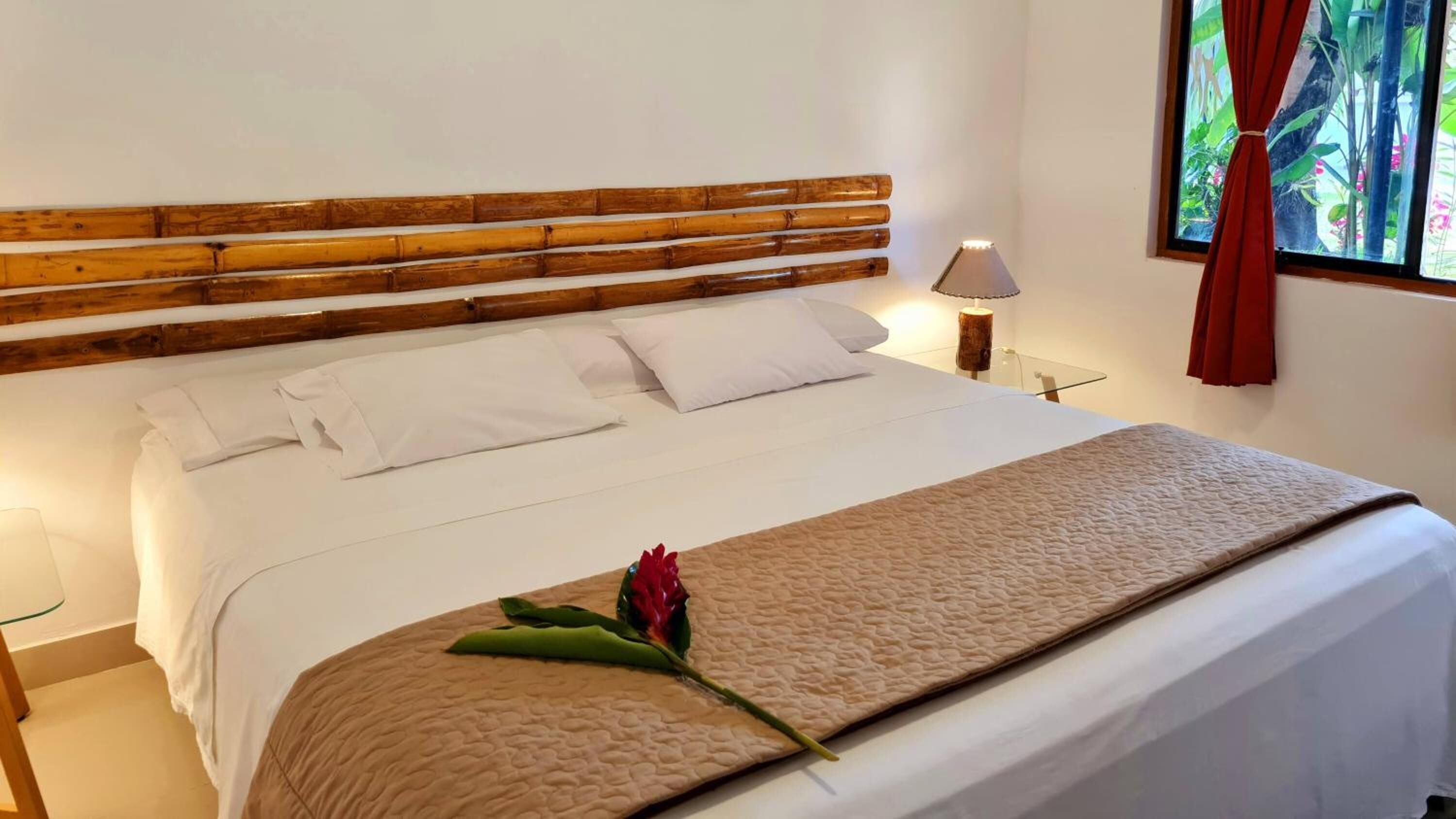 A neatly made bed with white linens and a beige quilted blanket, a wooden headboard, a bedside lamp, and a window with red curtains.