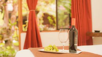 Una mesa con una botella de vino, una copa y un plato de uvas en una habitación con cortinas rojas y una ventana que da a follaje verde.