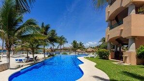 A swimming pool with lounge chairs, palm trees, and a building with balconies. - Listen to the ocean at night! (puerta aventuras)