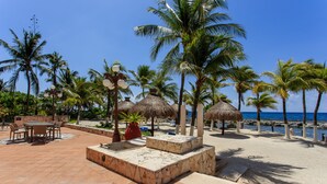 A beachside patio with a table and chairs, palm trees, and a thatched-roof structure. - Listen to the ocean at night! (puerta aventuras)