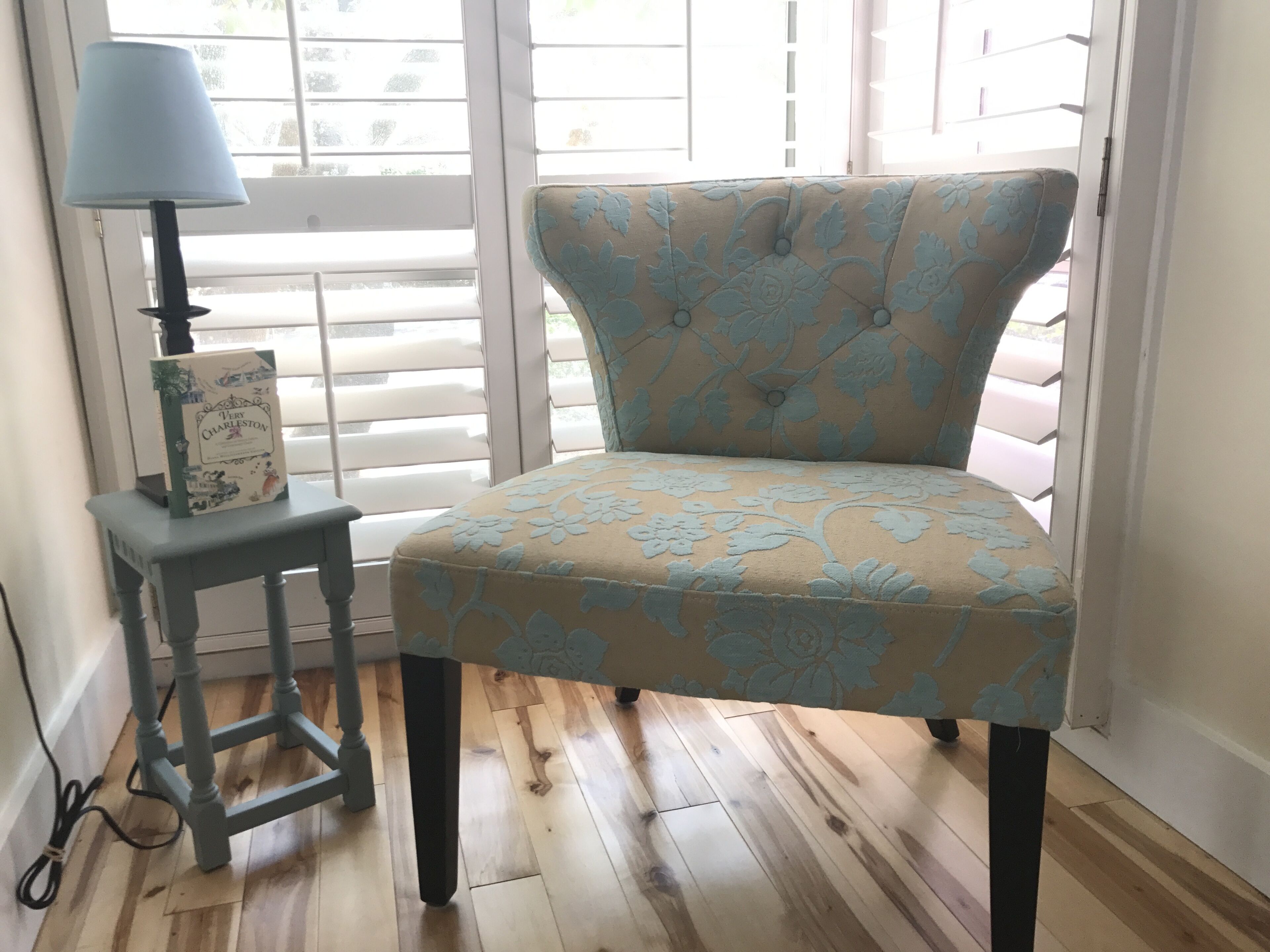 Een stoel met een bloemendesign, een bijzettafeltje met een lamp, en een boek getiteld 'Very Charleston' op het tafeltje.