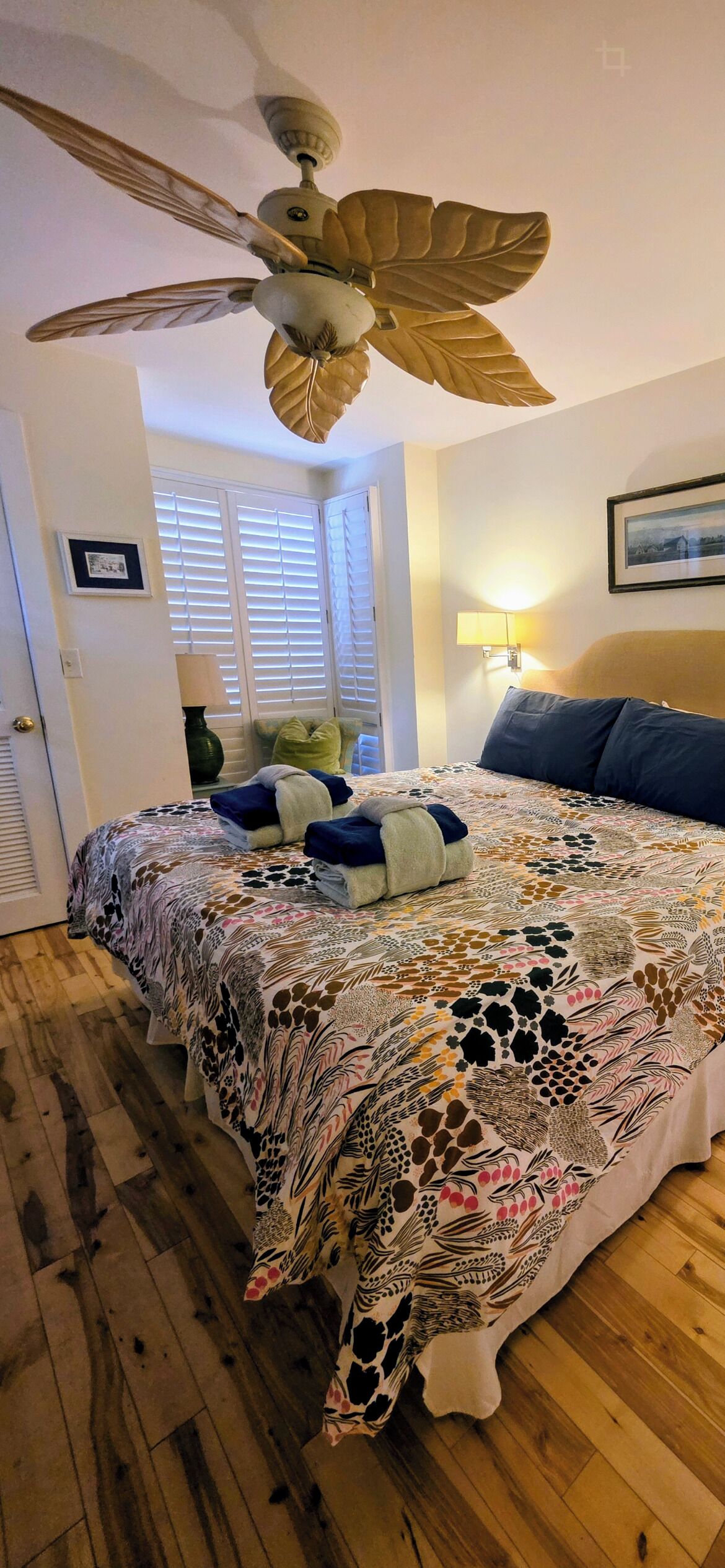 A bedroom with a large bed, a ceiling fan, a window with blinds, a bedside lamp, and a framed picture on the wall.