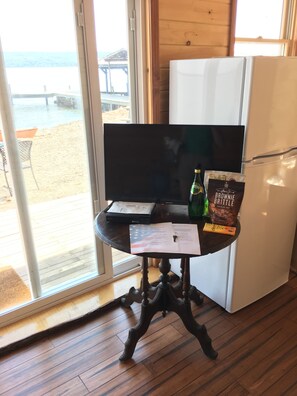A room with a wooden floor, a table with a flat-screen TV, a refrigerator, and a view of the beach through a glass door. - Great Cayuga Lake House Complex for families, reunions, or just relaxing! (Trumansburg)