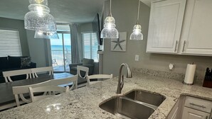 A kitchen with granite countertops, stainless steel sink, white cabinets, pendant lights, and a view to the outside. - Seawatch 2BR Oceanfront Wrap Around Balcony 916 ST (Myrtle Beach)