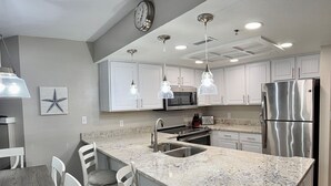 Une cuisine moderne avec des armoires blanches, des comptoirs en granit, des appareils électroménagers en acier inoxydable, des luminaires suspendus et une horloge au mur.