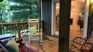 A wooden deck with a swing chair and a rocking chair, a wooden bench with cushions, and a view of the interior through an open door.