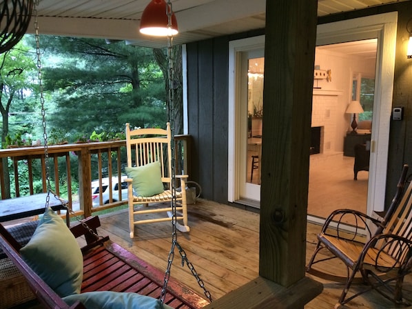 A wooden deck with a swing chair and a rocking chair, a wooden bench with cushions, and a view of the interior through an open door.