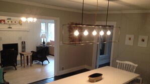 A modern kitchen with a long white countertop, a hanging light fixture with multiple bulbs, and a fireplace.