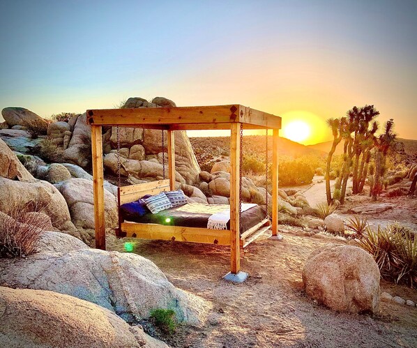 Estructura de cama de madera con asiento tipo columpio en un paisaje desértico con rocas y cactus.