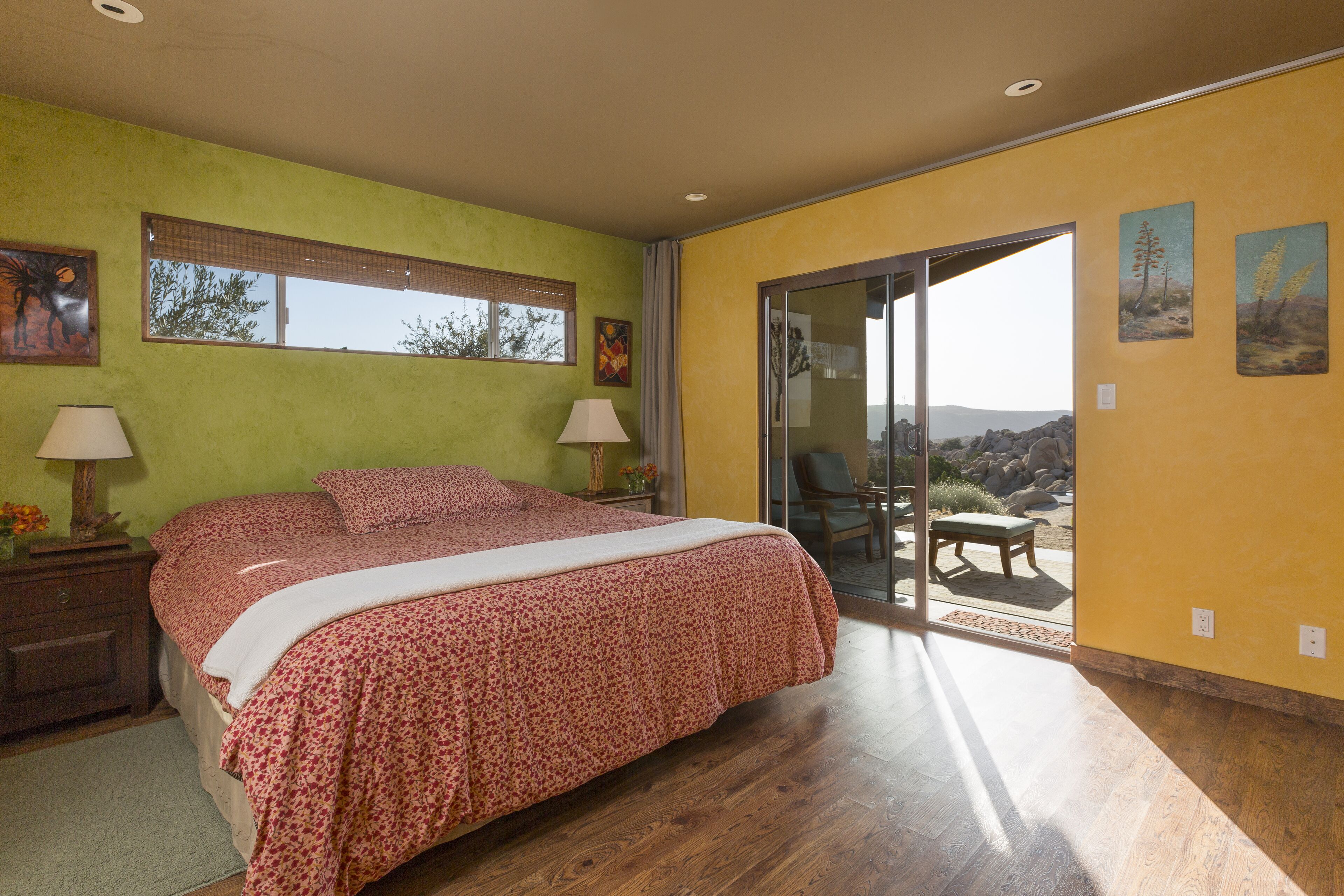 A bedroom with a bed, two bedside tables with lamps, a window with blinds, and a view of the outdoors.