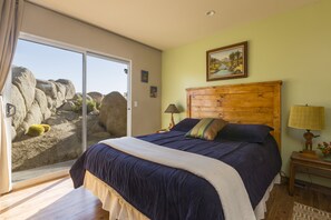 Un dormitorio con una cama grande, cabecera de madera, mesita de noche con lámpara y vistas a rocas a través de una puerta corrediza de vidrio.
