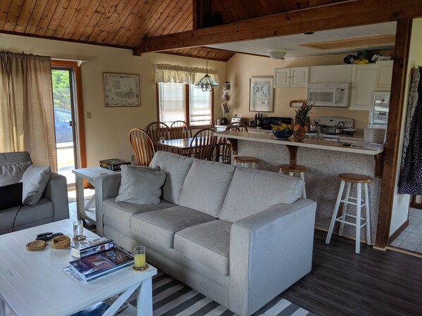 Uma sala de estar acolhedora com um sofá cinzento, uma mesa de centro com livros e um copo de suco, um espaço de jantar com cadeiras de madeira e uma cozinha com armários e eletrodomésticos brancos.