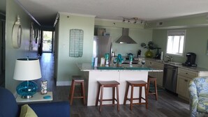 A modern kitchen with a central island, wooden stools, and a dining area with a blue sofa and a patterned armchair. - Ocean Front 1st Floor walkout to 25'x50' deck on the Boardwalk/Beach at 4th St (Ocean City)