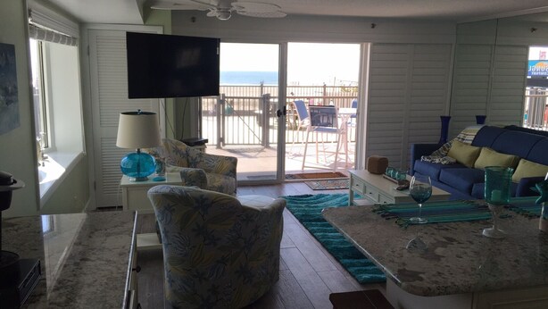 A spacious living area with a large TV, a sofa, a coffee table, and a view of the beach through the sliding glass door. - Ocean Front 1st Floor walkout to 25'x50' deck on the Boardwalk/Beach at 4th St (Ocean City)