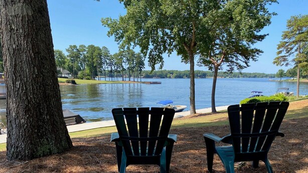 Two Adirondack chairs facing a lake with a dock and boats, surrounded by trees and a grassy area.