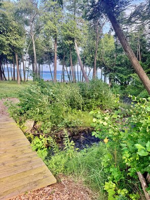 A wooden deck leading to a natural area with dense greenery and a view of the water. - Waterfront Condo On Little Traverse Bay. Beaches, Tennis,Near Golf And Ski (Harbor Springs)