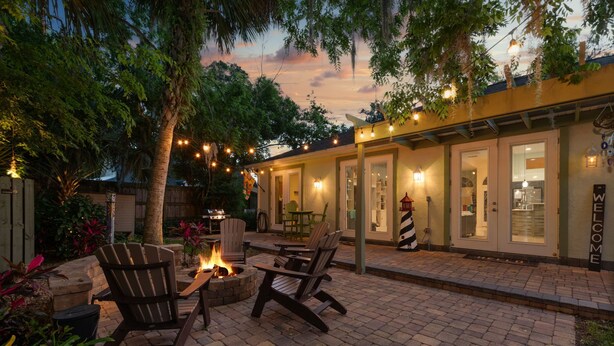 Un patio al aire libre con suelo de ladrillo, sillas de madera, un hogar para fuego y luces colgantes.