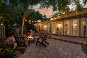 Un patio al aire libre con suelo de ladrillo, sillas de madera, un hogar para fuego y luces colgantes.