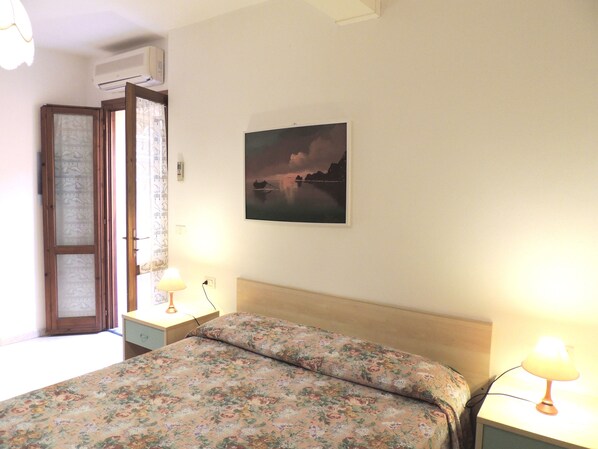 A bedroom with a bed, two bedside tables, a nightstand, a wall-mounted air conditioner, and a framed picture on the wall.