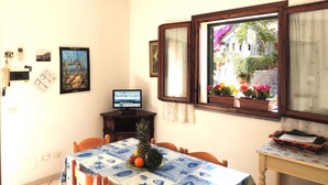A dining area with a table set for two, a computer on a desk, and a window with a view of the street and plants outside. - Appartamenti Montecristo e Pianosa (Campo nell'Elba)