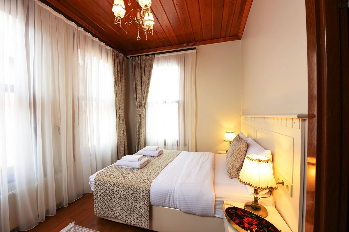 Une chambre d’hôtel avec un plafond en bois, un lit recouvert de draps blancs, une lampe de chevet et des rideaux en voile.