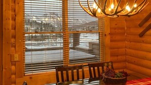 A wooden cabin interior with a dining table, chairs, a chandelier, and large windows.