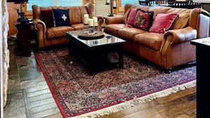 A spacious living room with a large leather sofa, a coffee table, and a patterned rug. There are wooden walls and a ceiling fan.